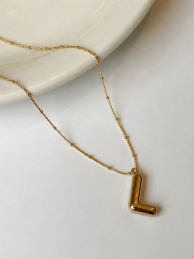 Découvrez le Collier Initiale L Élégance™. Une chaîne perlée dorée raffinée avec pendentif lettre L au design minimaliste et intemporel. Le cadeau parfait pour elle.
